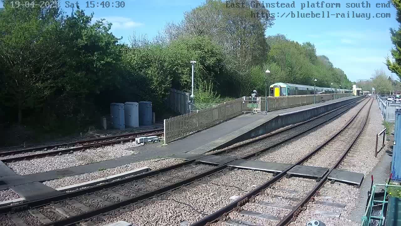 A train sits at a station platform on a sunny day, with trees and a fence lining the tracks.