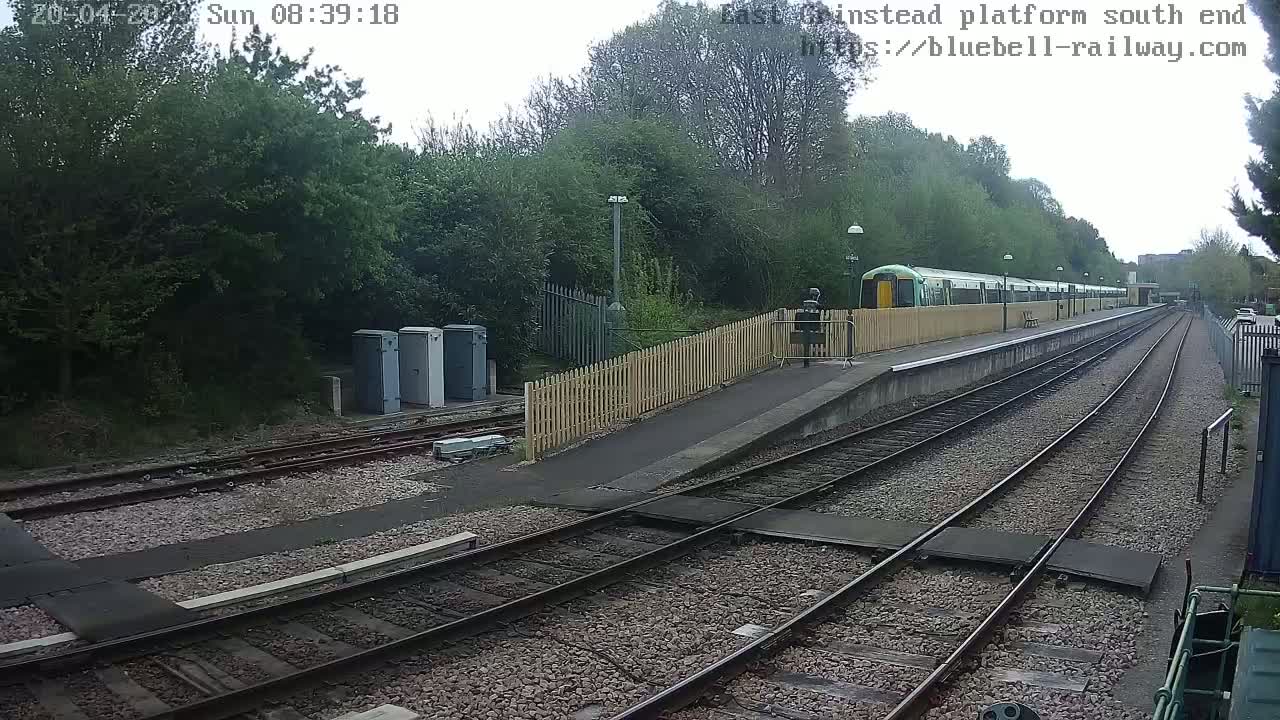 A train is at a station platform on an overcast day, with multiple train tracks visible extending into the distance.