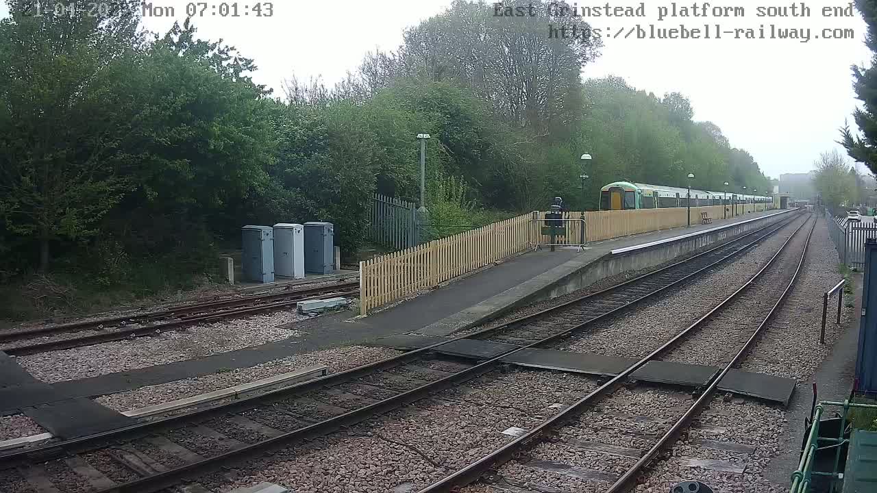 A train sits at a station platform on an overcast day, with multiple sets of train tracks extending into the distance, bordered by trees and a wooden fence.