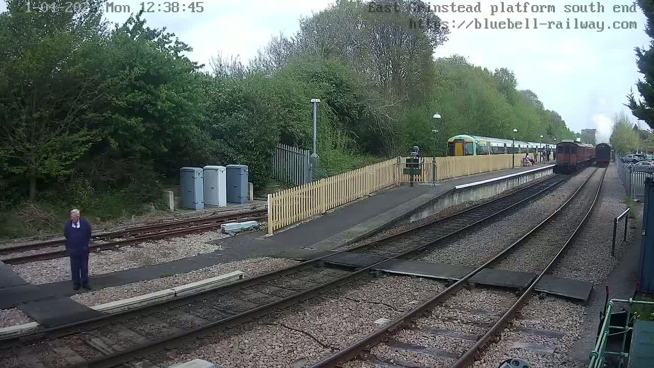 A train departs a railway station platform on an overcast day, past a person standing on the platform.
