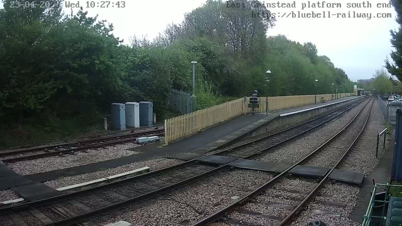 A view of a train platform on an overcast day, showing train tracks extending into the distance, bordered by a wooden fence and lush green trees.