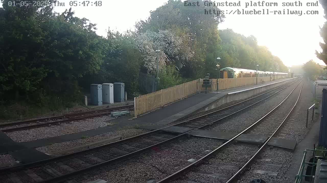 A train sits at a station platform on a sunny morning, surrounded by trees and a wooden fence.