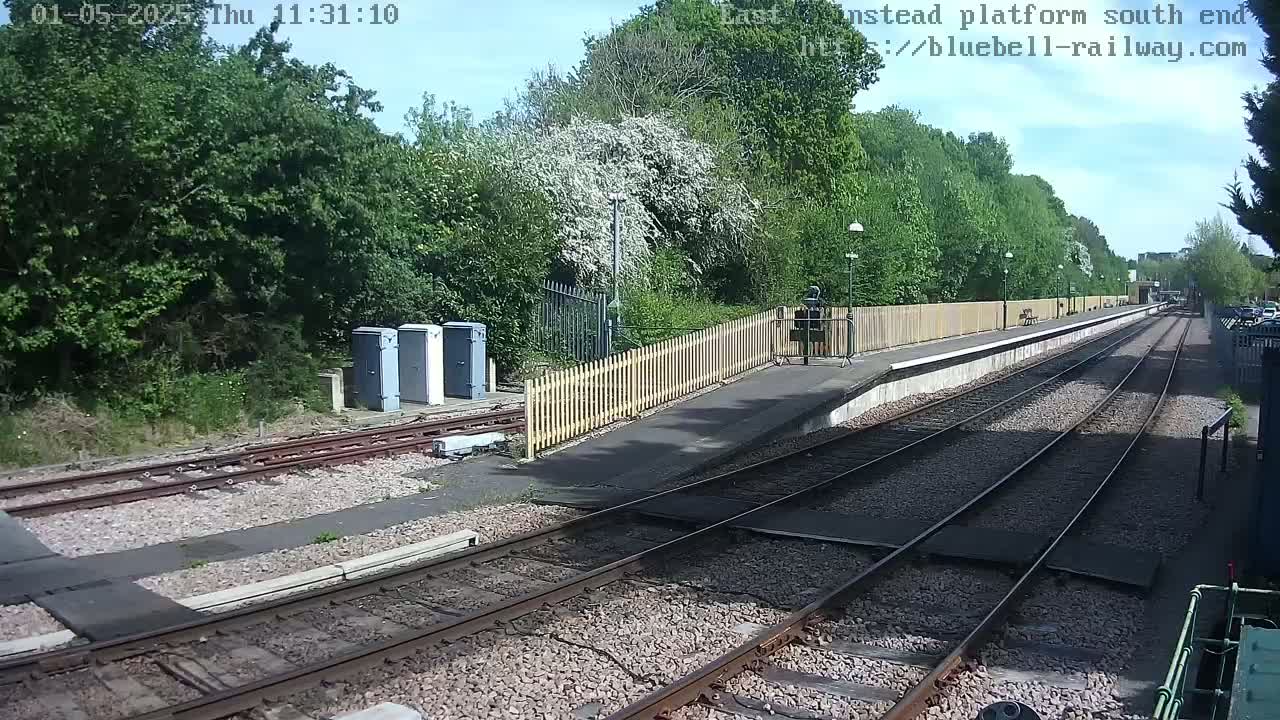 A train platform with a wooden fence and trees under a sunny sky extends into the distance along railway tracks.