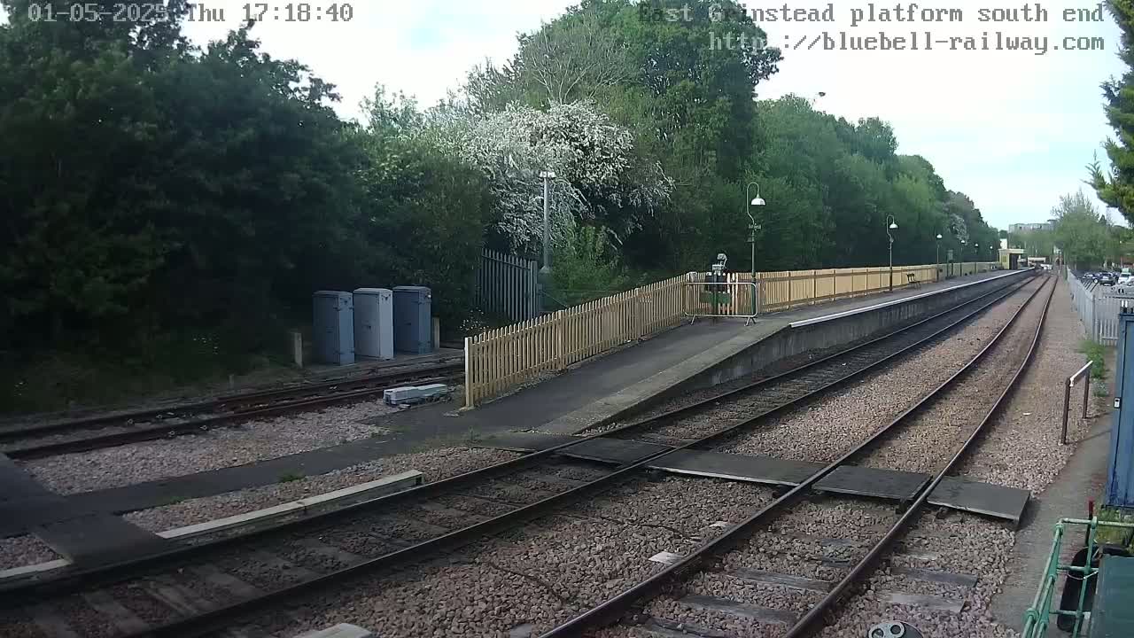 A railway platform with two sets of tracks, a wooden fence, and some trees, under a partly cloudy sky.
