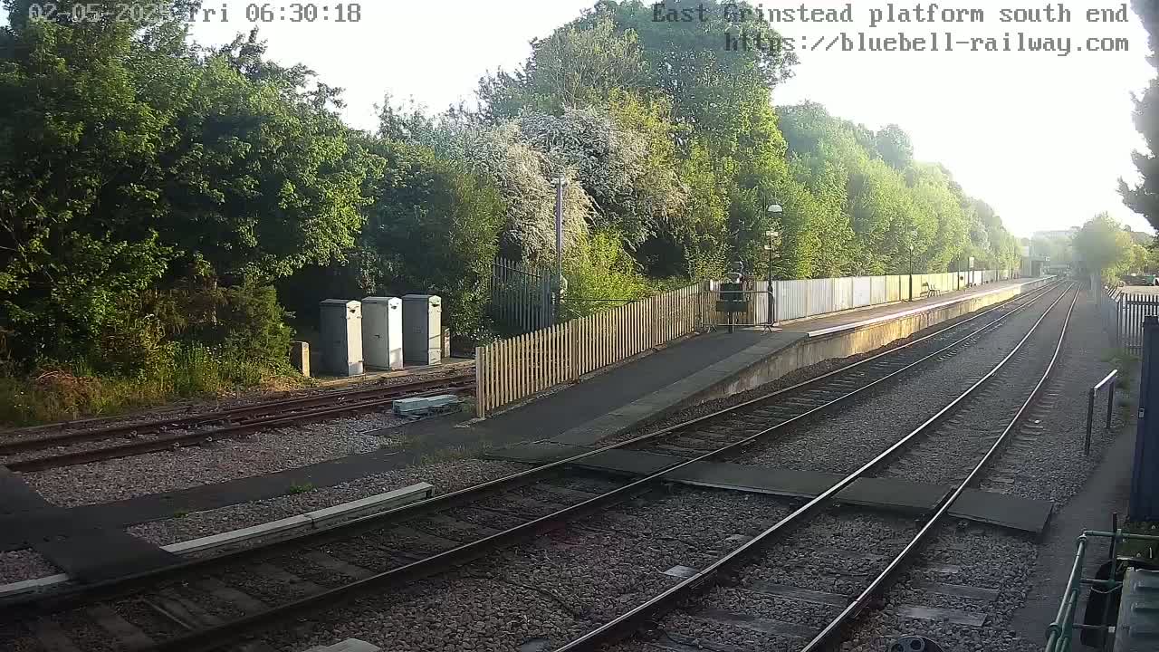 A sunny view of a train platform with two sets of tracks extending into the distance, bordered by a wooden fence and lush green trees.