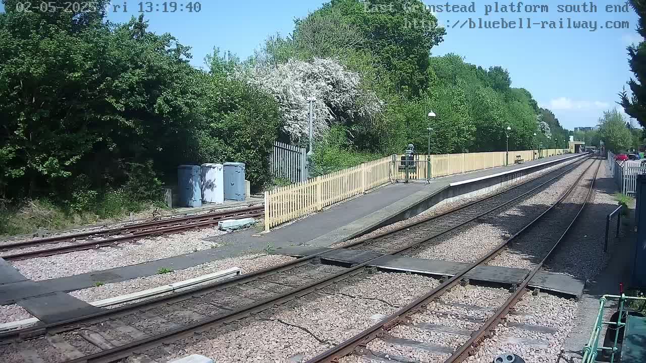 A train platform with two sets of tracks extends into the distance, bordered by a wooden fence and lush green trees under a mostly sunny sky.