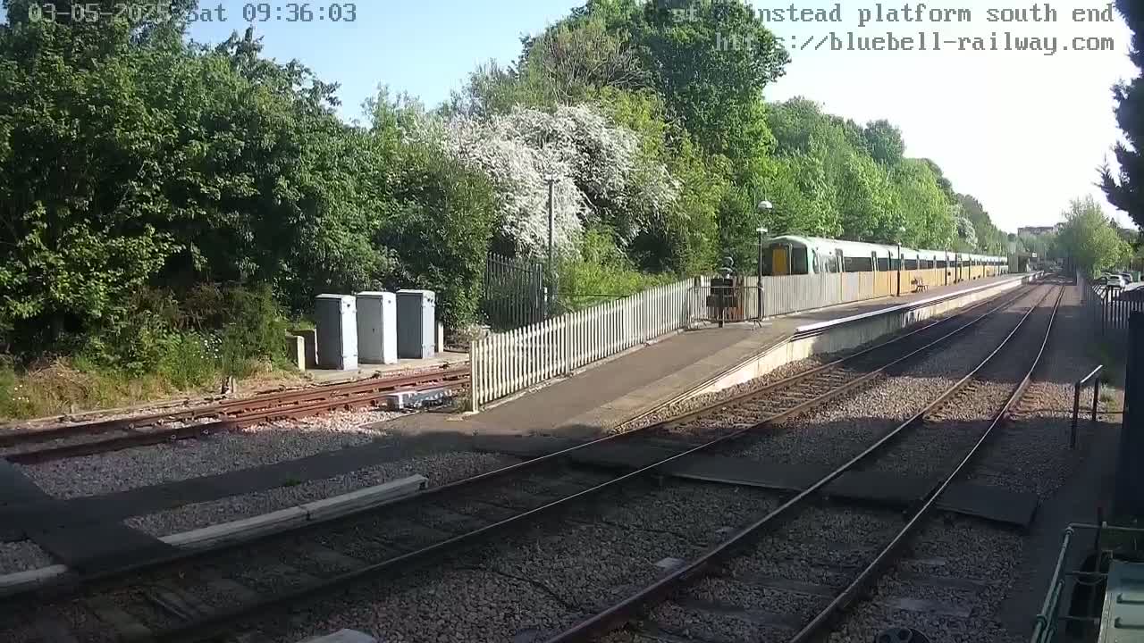 A sunny day at a train station shows a light-green train stopped at a platform next to a white picket fence and lush green trees.