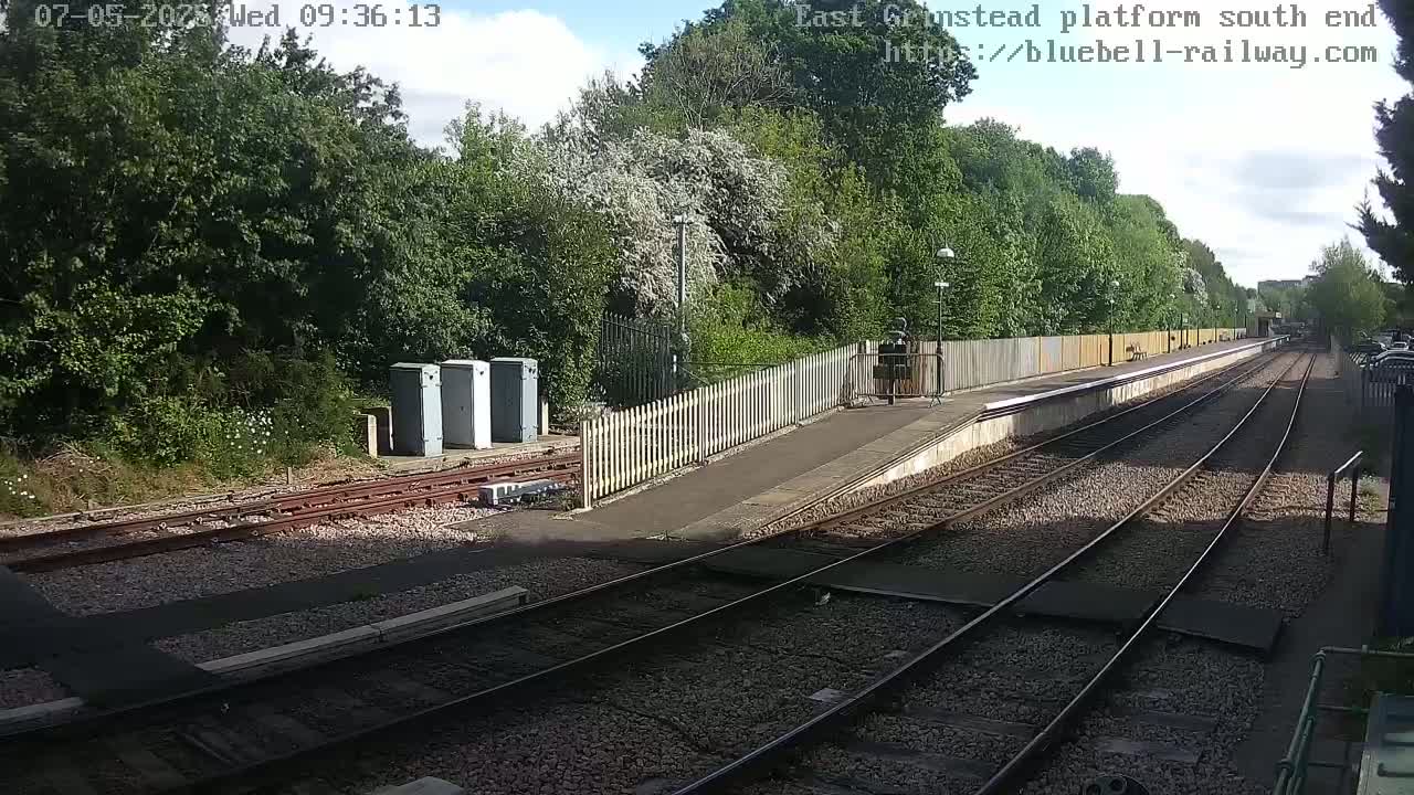 A partly sunny view of a train platform bordered by trees and a wooden fence, with railway tracks extending into the distance.