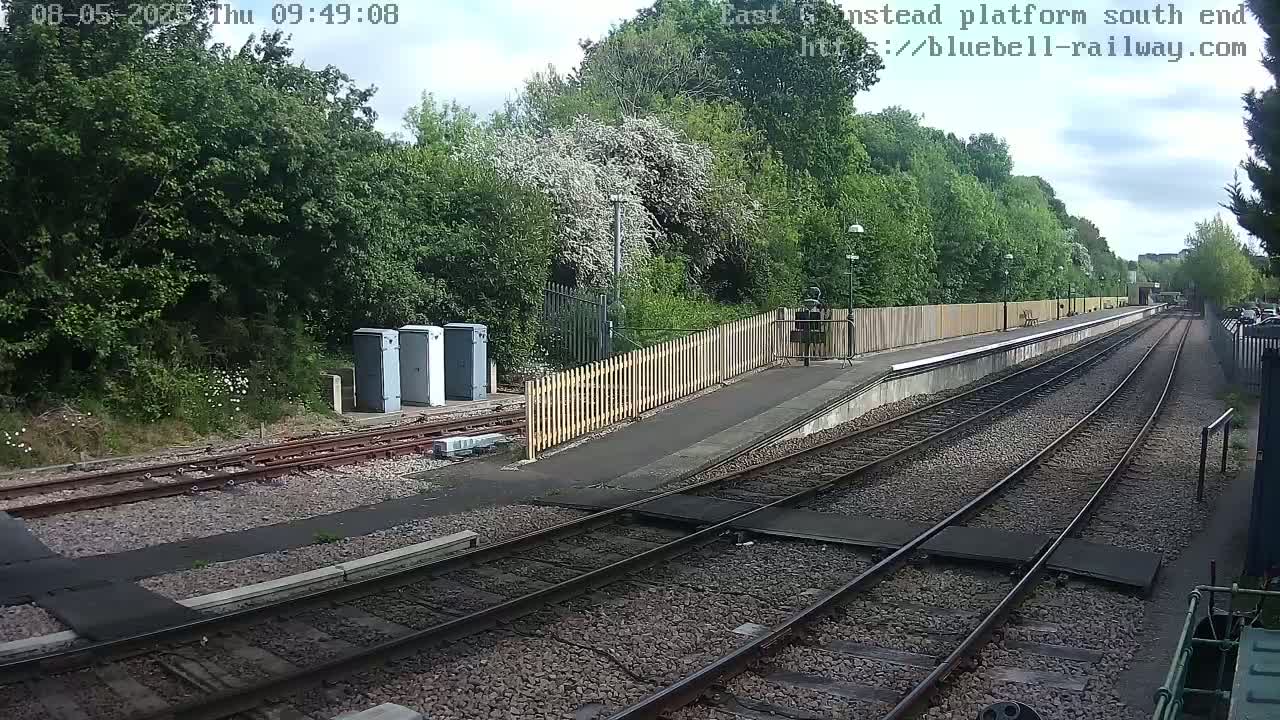 A partly cloudy day at a train station with a wooden fence and platform next to a lush green wooded area.