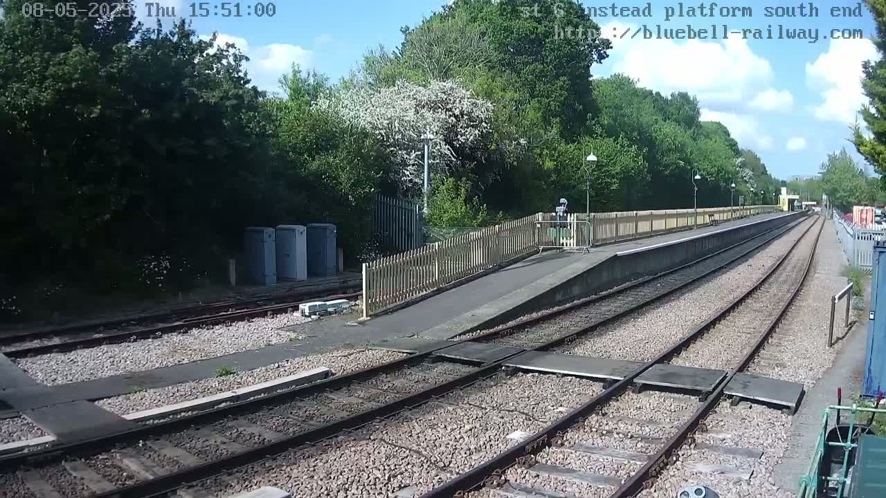 A sunny day at a train station with a platform, tracks, and lush green trees surrounding the area.
