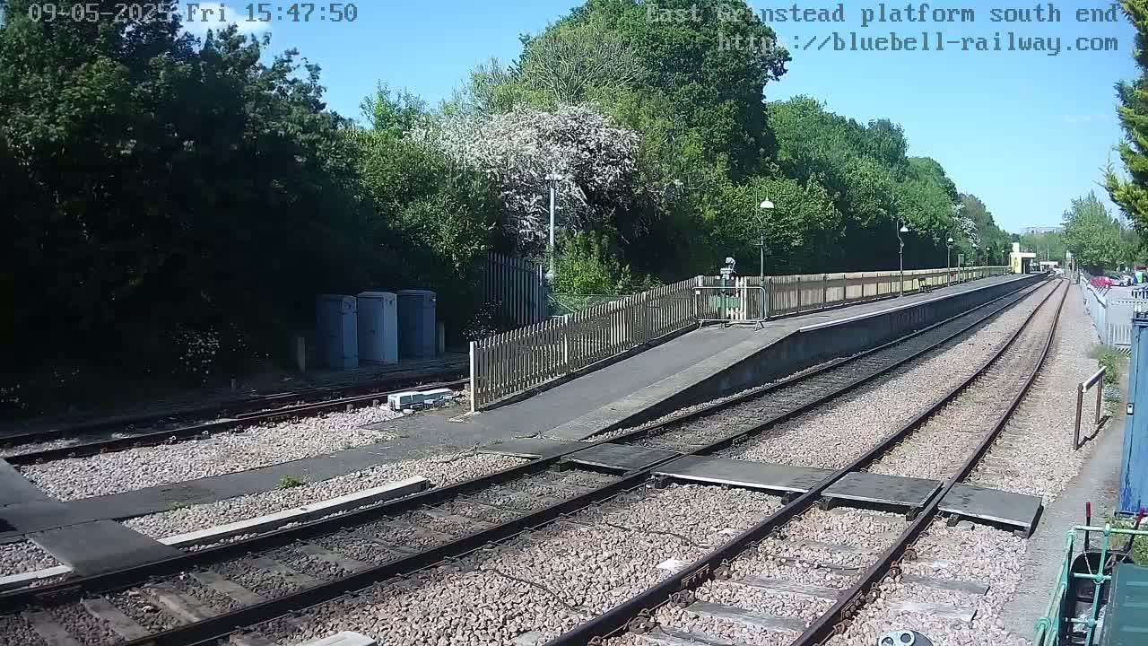 A sunny view of a train platform with multiple tracks, a wooden fence, and lush green trees in the background.