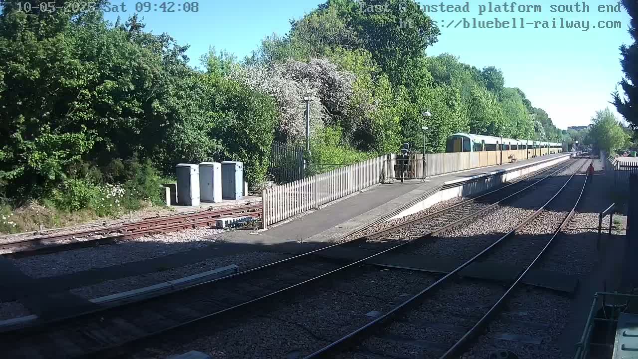A sunny day at a train station shows a train at a platform, railway tracks extending into the distance, and lush green trees and vegetation surrounding the area.