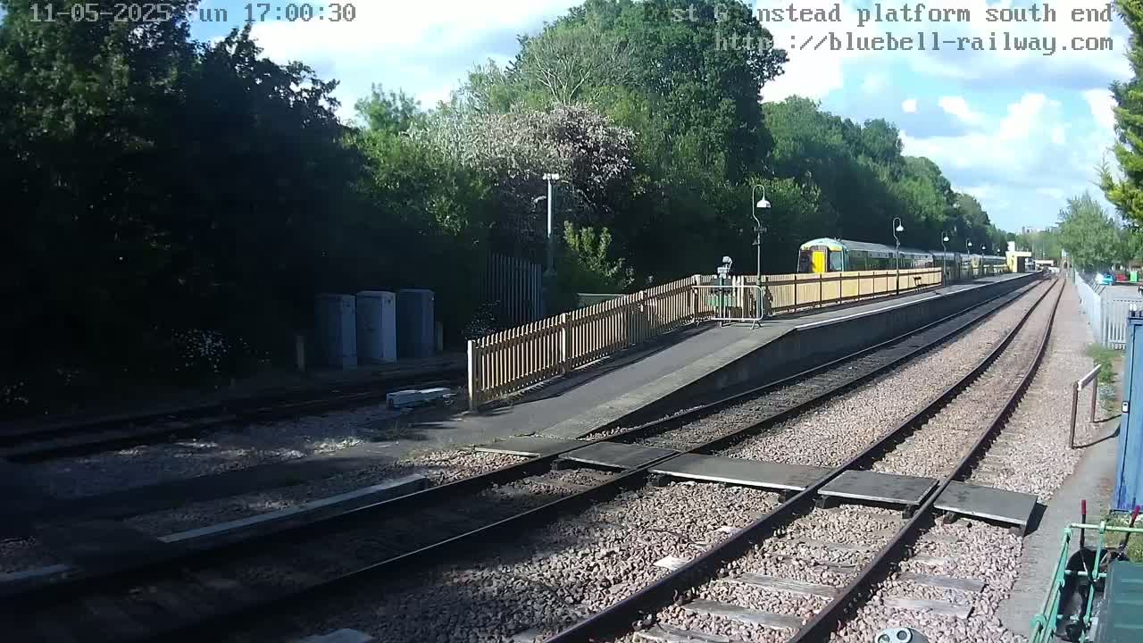 A partly sunny day at a train station shows a passenger train at the platform, alongside train tracks that extend into the distance, bordered by a wooden fence and lush green trees.