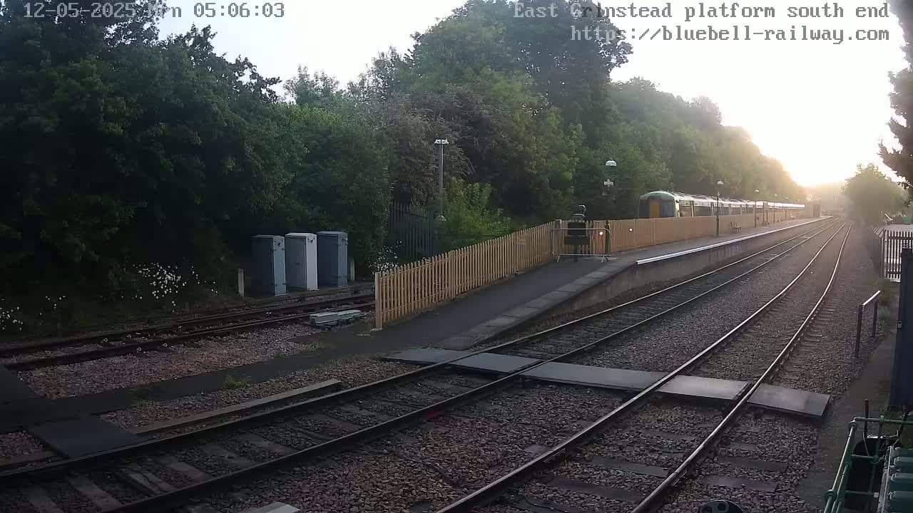A train sits at a station platform on a sunny morning, with lush green trees lining the railway tracks.