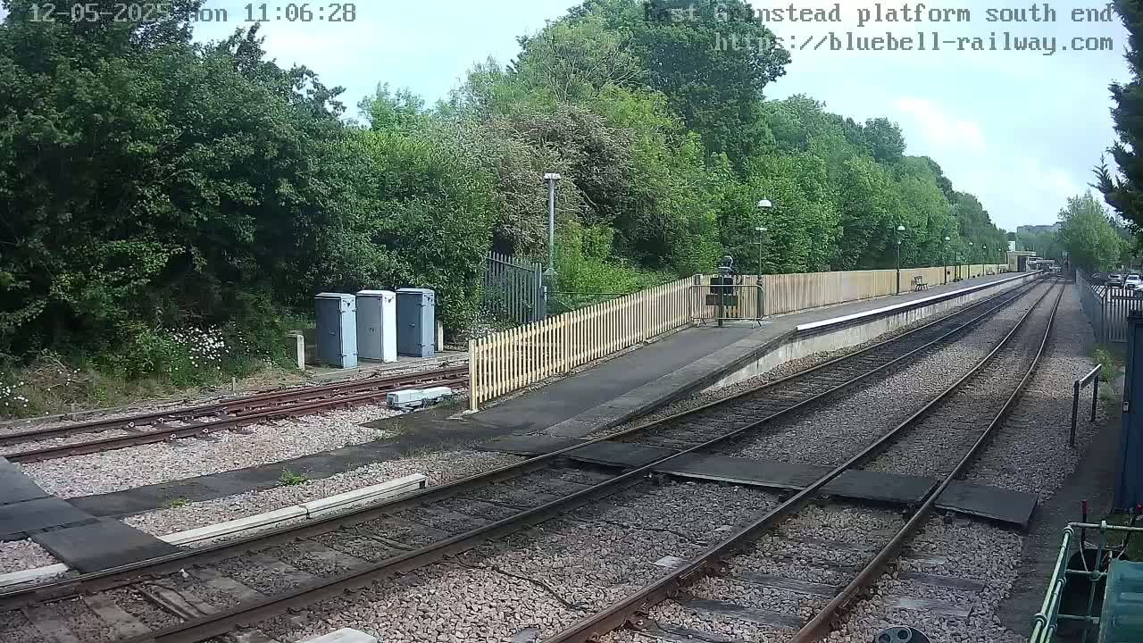 A train platform with a wooden fence and lush green trees under a partly cloudy sky.