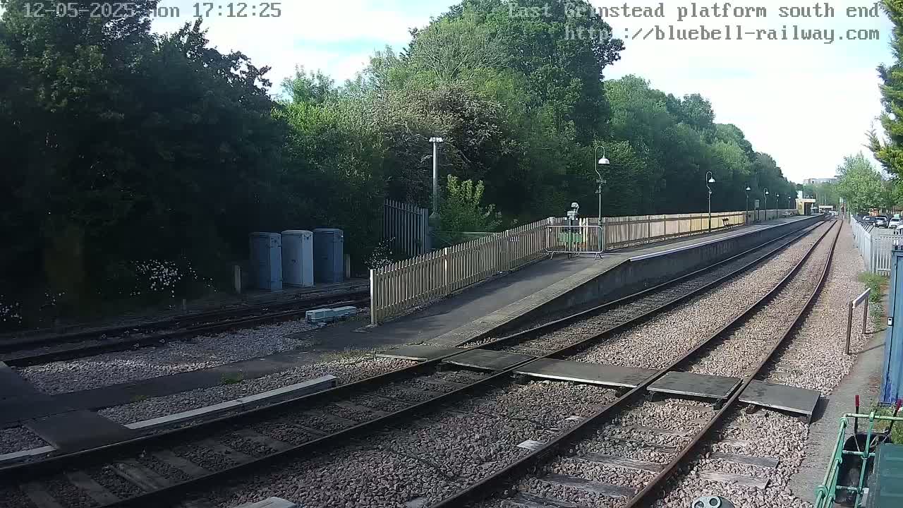 A mostly sunny view of a train platform with two sets of tracks and a wooden fence, surrounded by lush green trees.