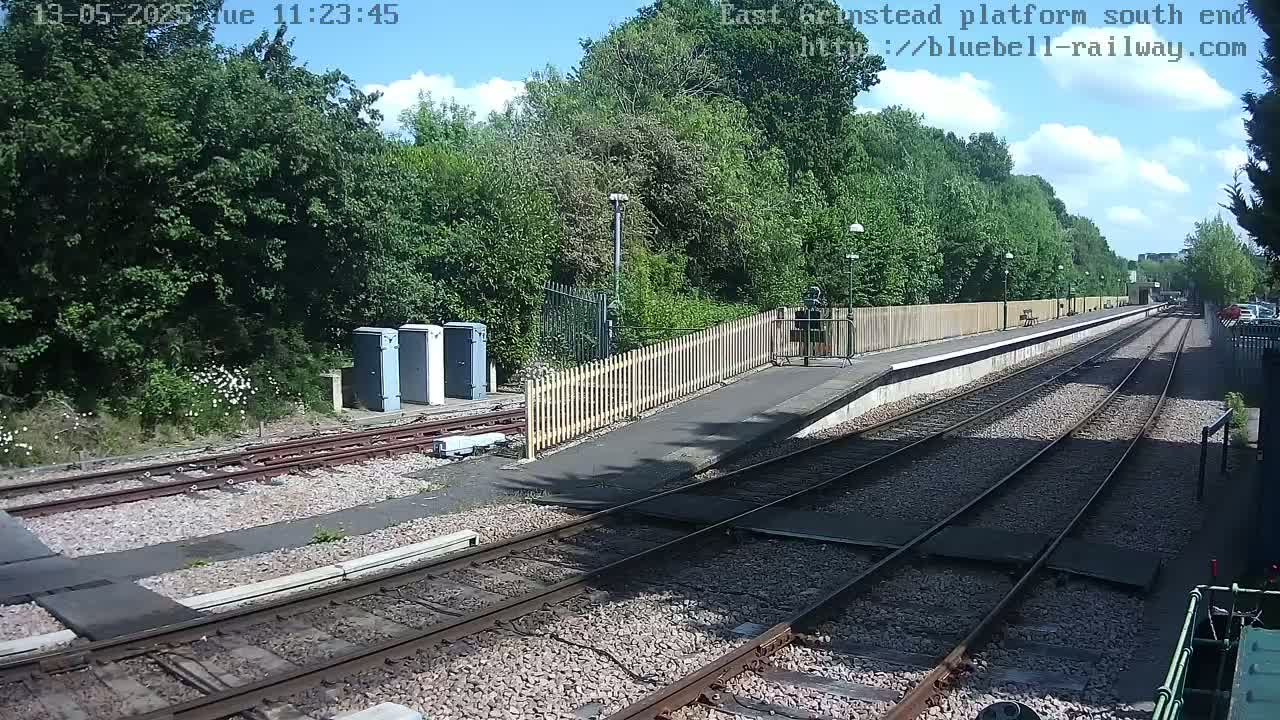 A sunny day at a train station platform, showing railway tracks, a platform with a wooden fence, and lush green trees in the background.