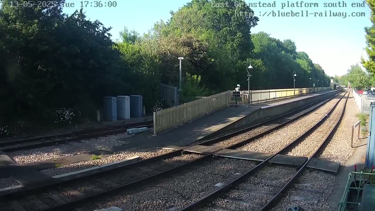 A sunny view of a train platform with multiple tracks and a wooden fence, surrounded by lush greenery.