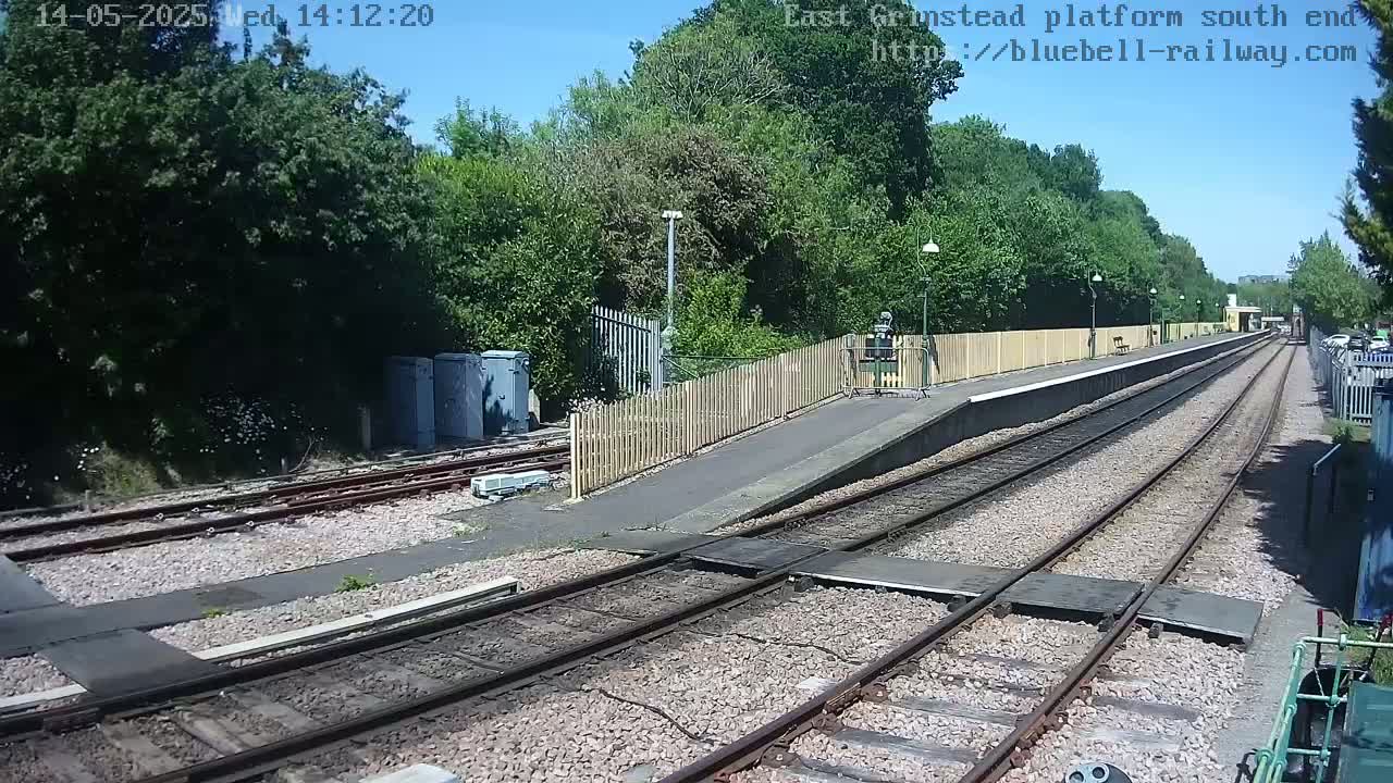 A sunny view of a train platform with two sets of tracks extending into the distance, surrounded by lush green trees and a wooden fence.