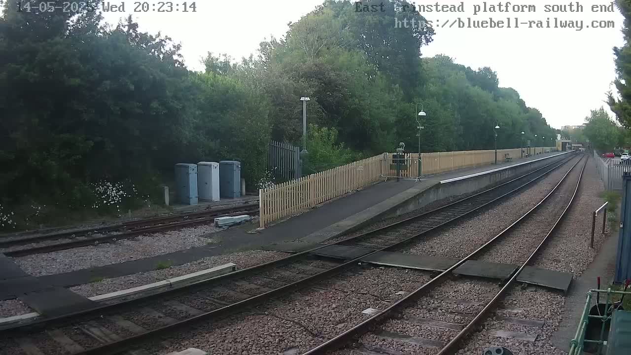 A train platform and railway tracks are visible, surrounded by lush green trees and shrubs under overcast skies.