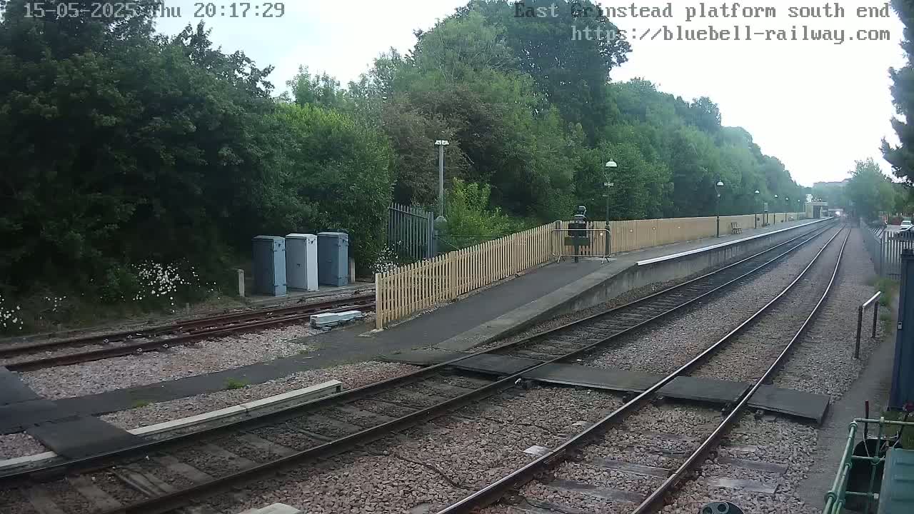 A train platform with two sets of tracks extends into the distance under an overcast sky, bordered by lush green vegetation and a wooden fence.
