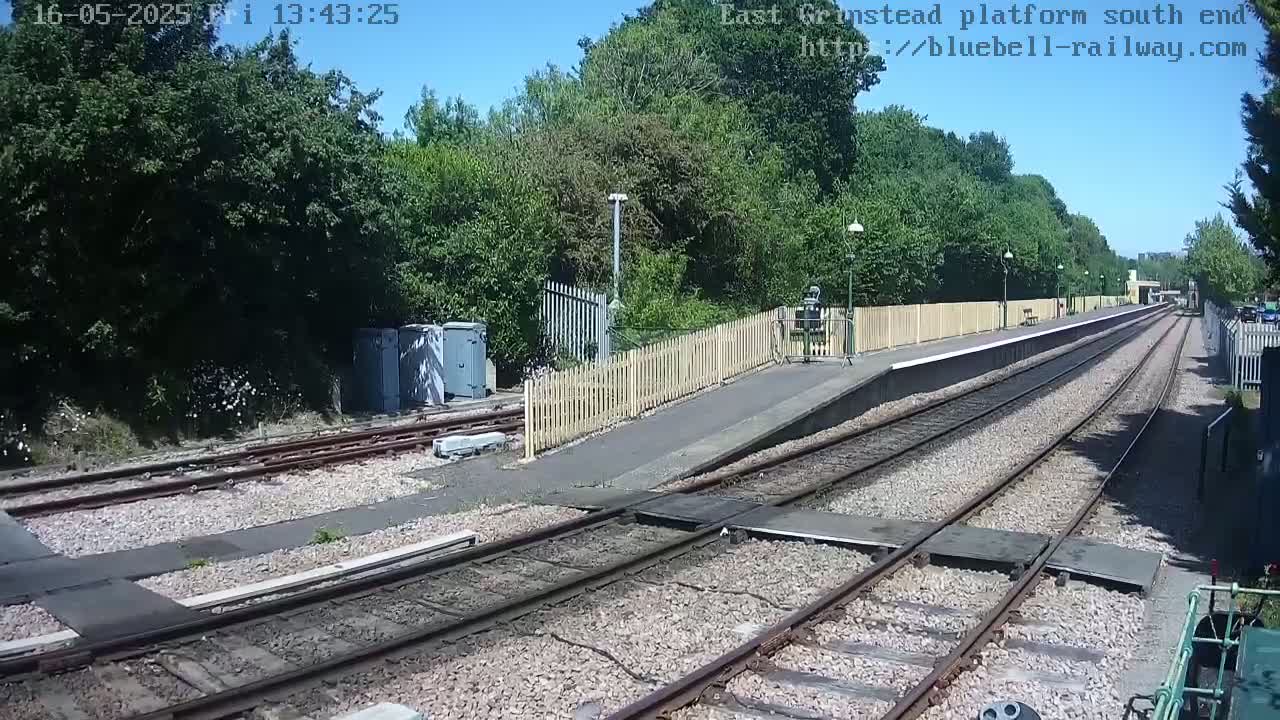 A sunny day at a train platform with a wooden fence, railway tracks, and lush green trees surrounding the area.