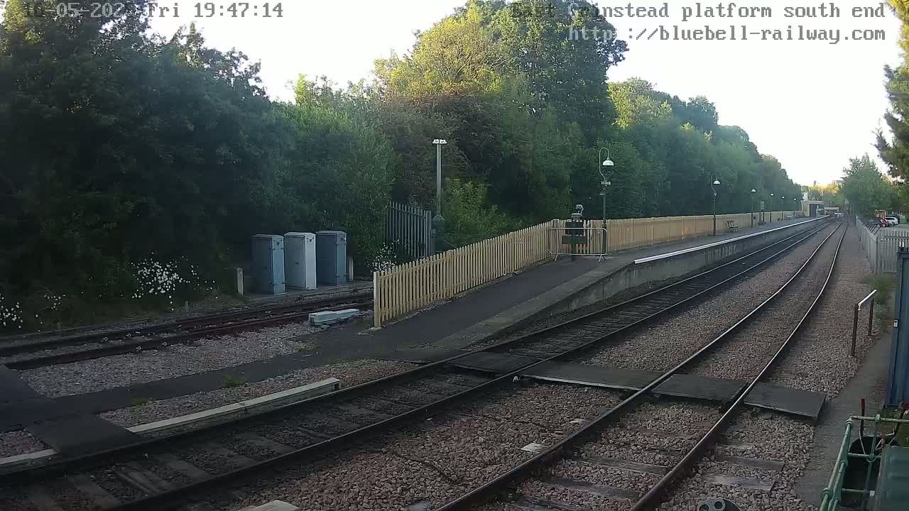 A train station platform with a wooden fence, several railway tracks, and lush green trees under a partly sunny sky.