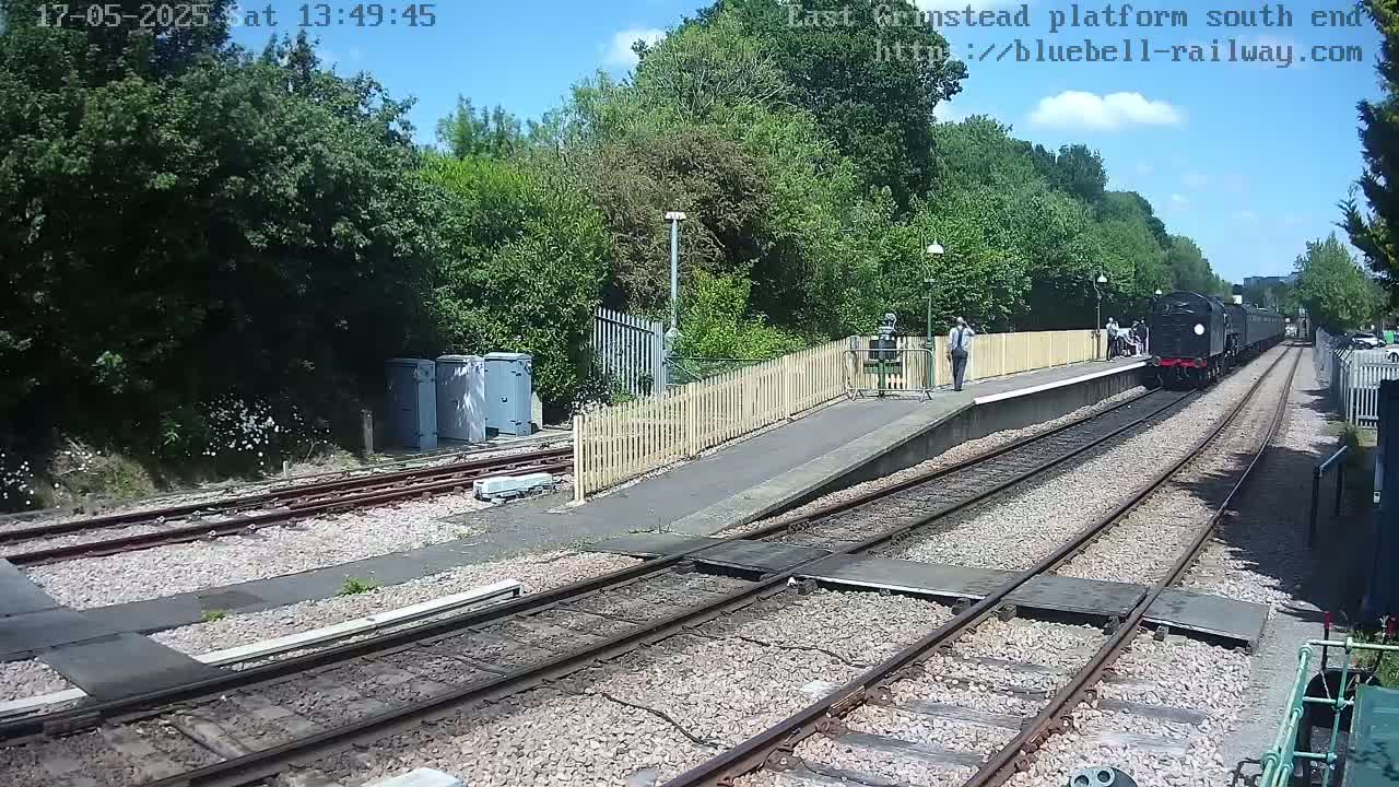 A black steam train departs from a railway station platform on a sunny day, passing alongside a wooden fence and lush green trees.