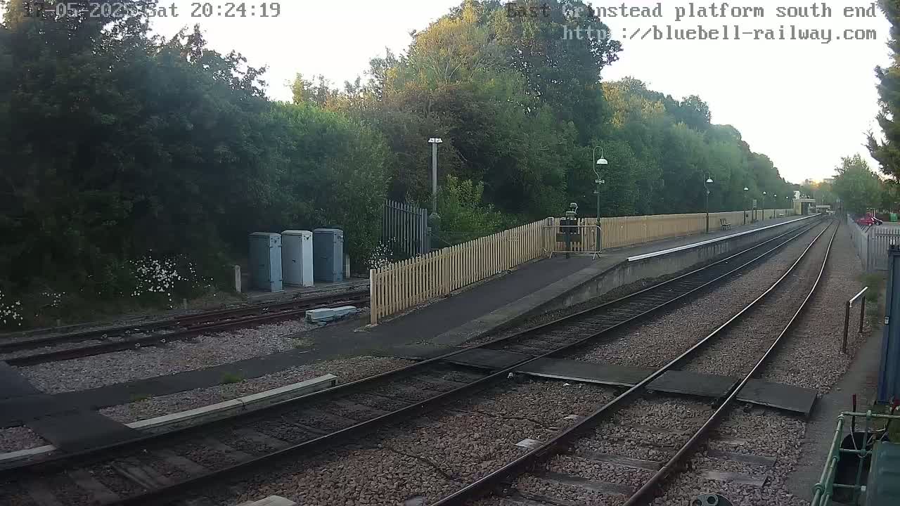 A view of a train platform at dusk, showing multiple train tracks extending into the distance, surrounded by lush green trees and a wooden fence.
