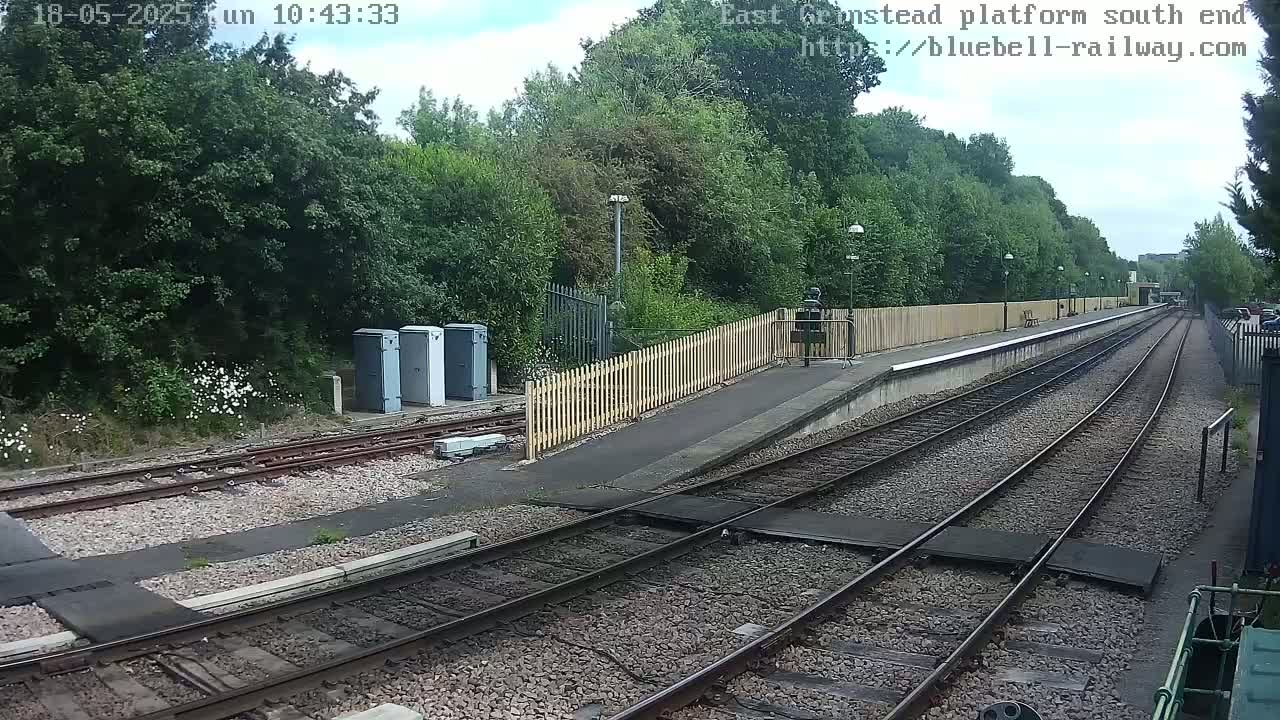 A view of a train platform on a partly cloudy day, showing train tracks extending into the distance, bordered by a wooden fence and lush green trees.