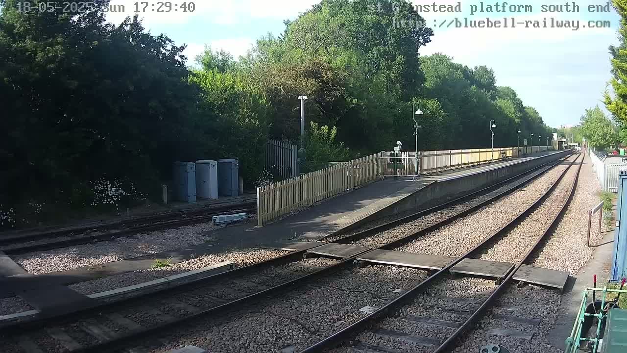 A mostly sunny view of a train station platform with multiple train tracks and a wooden fence, surrounded by lush green trees.