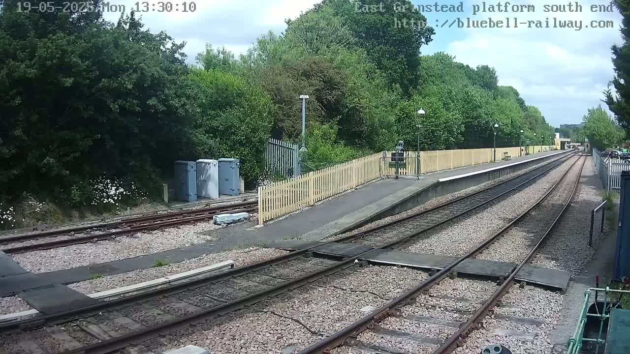A view of a train platform with two sets of tracks extending into the distance on a partly cloudy day, surrounded by lush green vegetation.