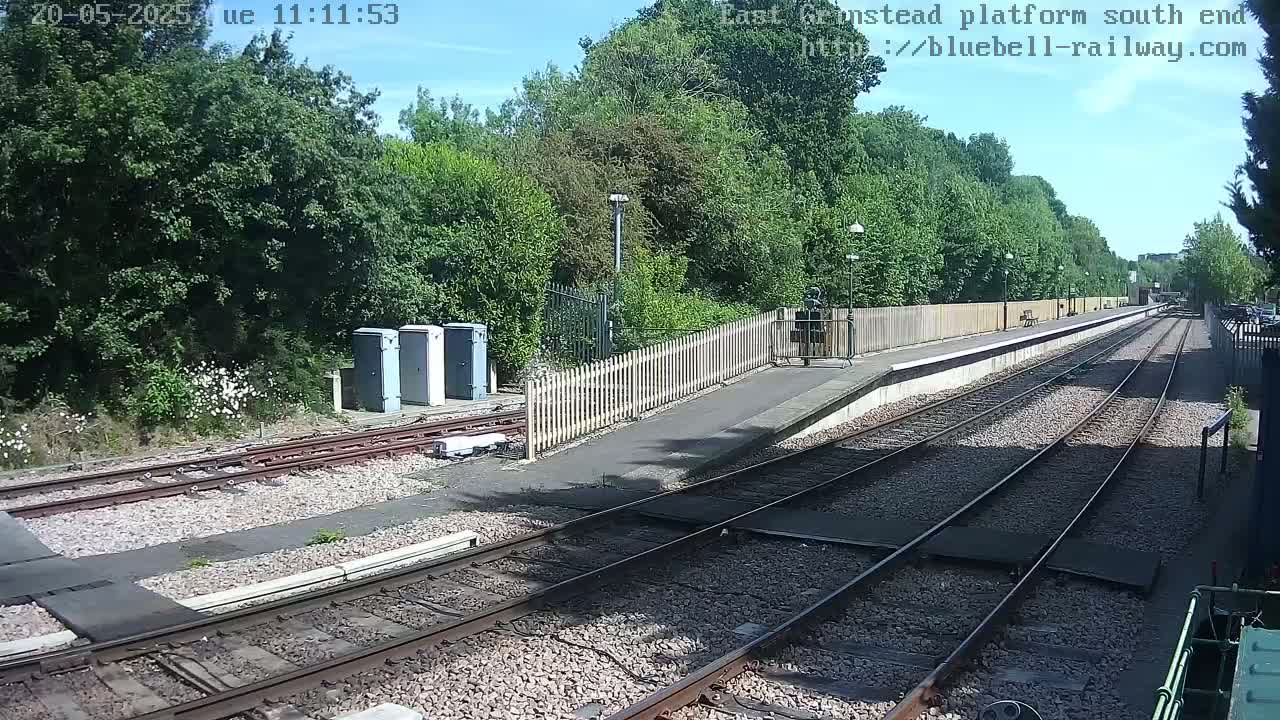 A sunny day view of a train platform with two sets of tracks extending into the distance, bordered by a wooden fence and lush green trees.
