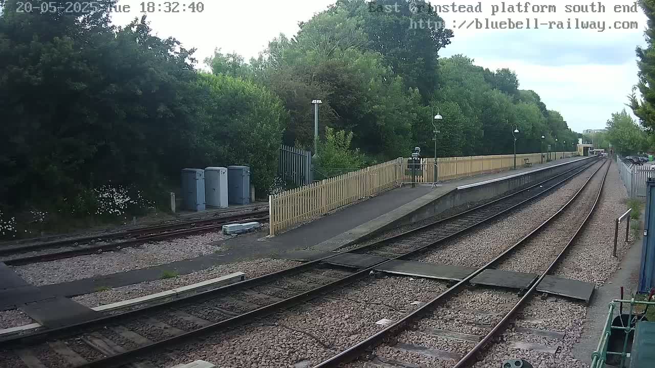 A view of a train platform with two sets of tracks, bordered by a wooden fence and lush green trees under an overcast sky.