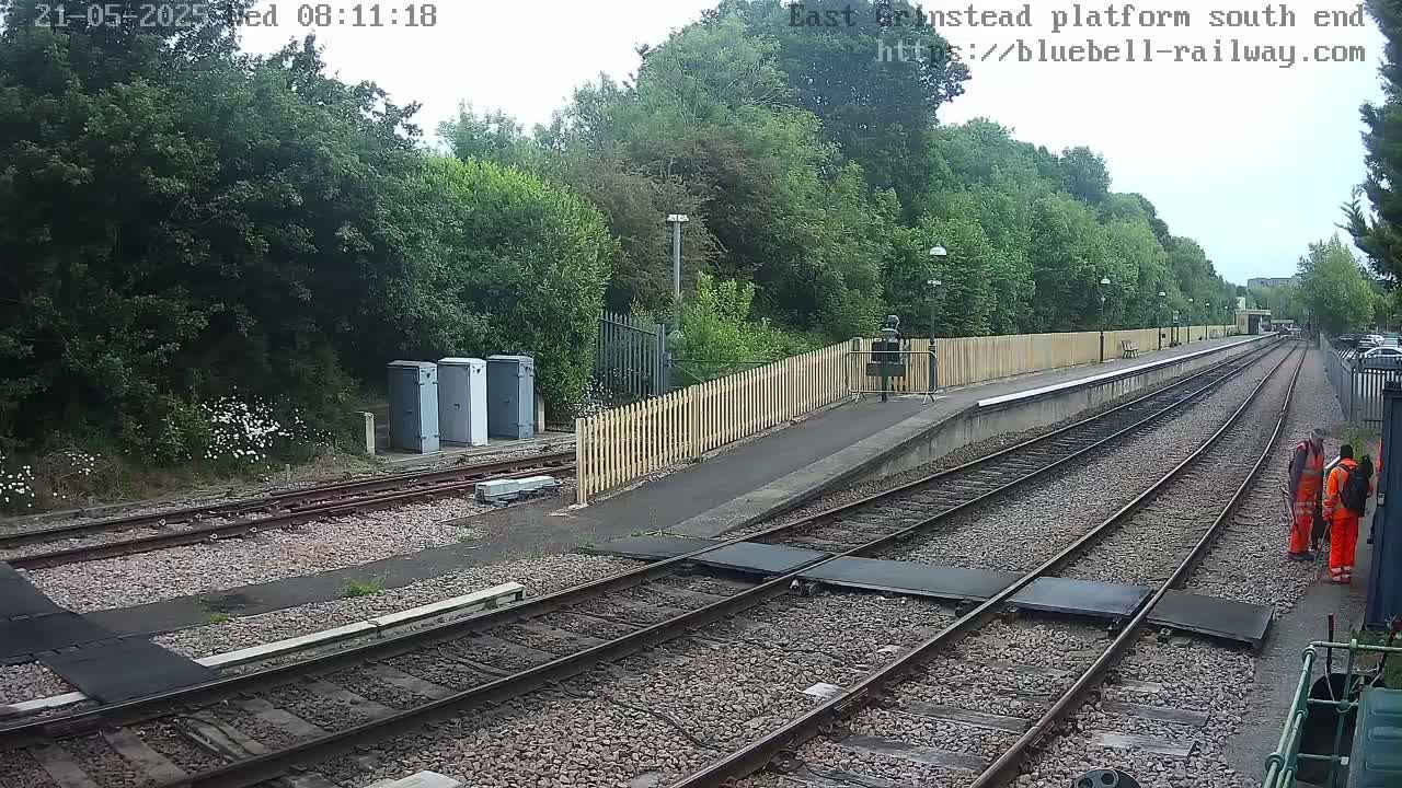 A train platform with two workers standing nearby, under an overcast sky, bordered by trees and a wooden fence.