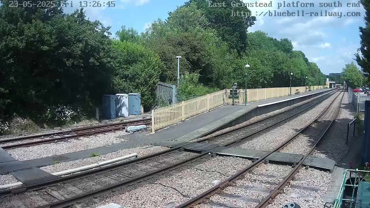 A sunny day at a train station with a view down the railway tracks, surrounded by lush green trees and shrubbery.