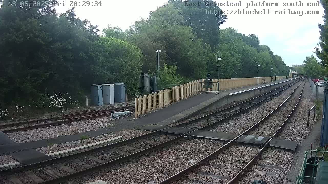 A train platform with two sets of tracks extends into the distance, bordered by a wooden fence and lush green trees under a partly cloudy sky.