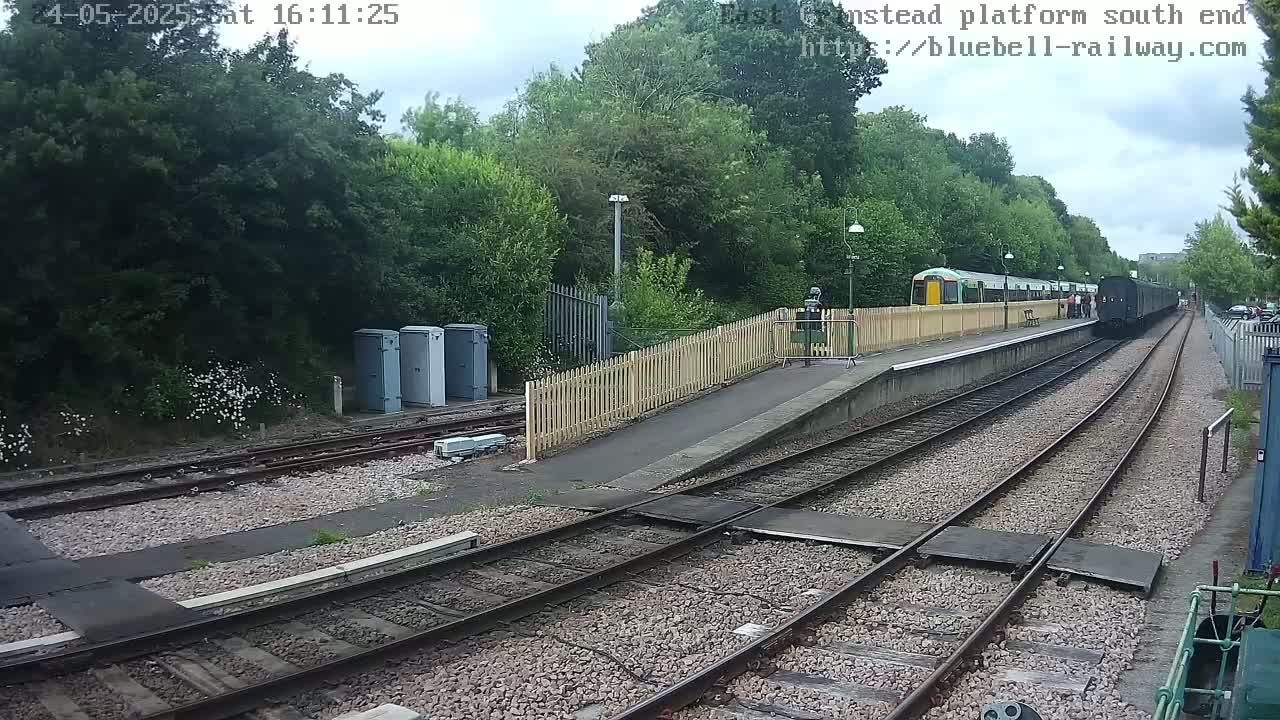 A train departs from a railway station platform on an overcast day, surrounded by lush green vegetation.