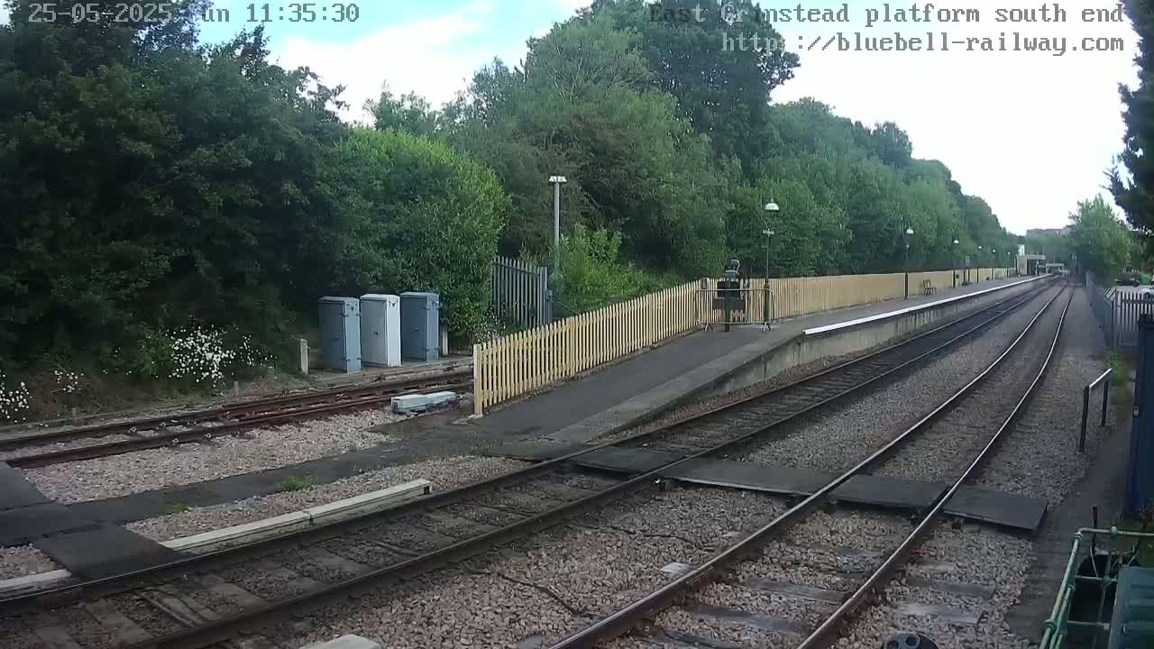 A mostly sunny view of a train platform with two sets of tracks, a wooden fence, and lush green vegetation behind it.