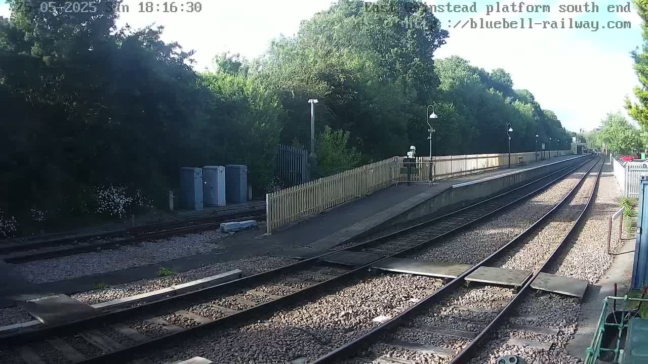 A train platform with two sets of tracks extends into the distance, surrounded by lush green trees under a bright, sunny sky.