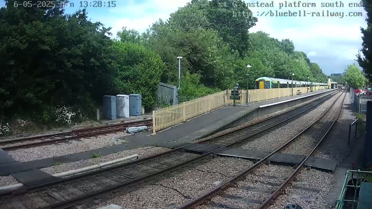 A train sits at a station platform on a partly sunny day, with lush green trees and vegetation surrounding the railway tracks.