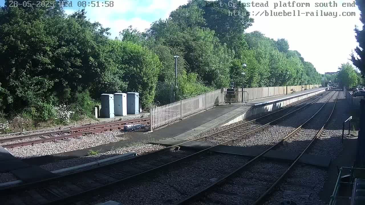 A mostly sunny view of a train platform with several train tracks, bordered by green trees and a wooden fence, with a few utility boxes near the tracks.