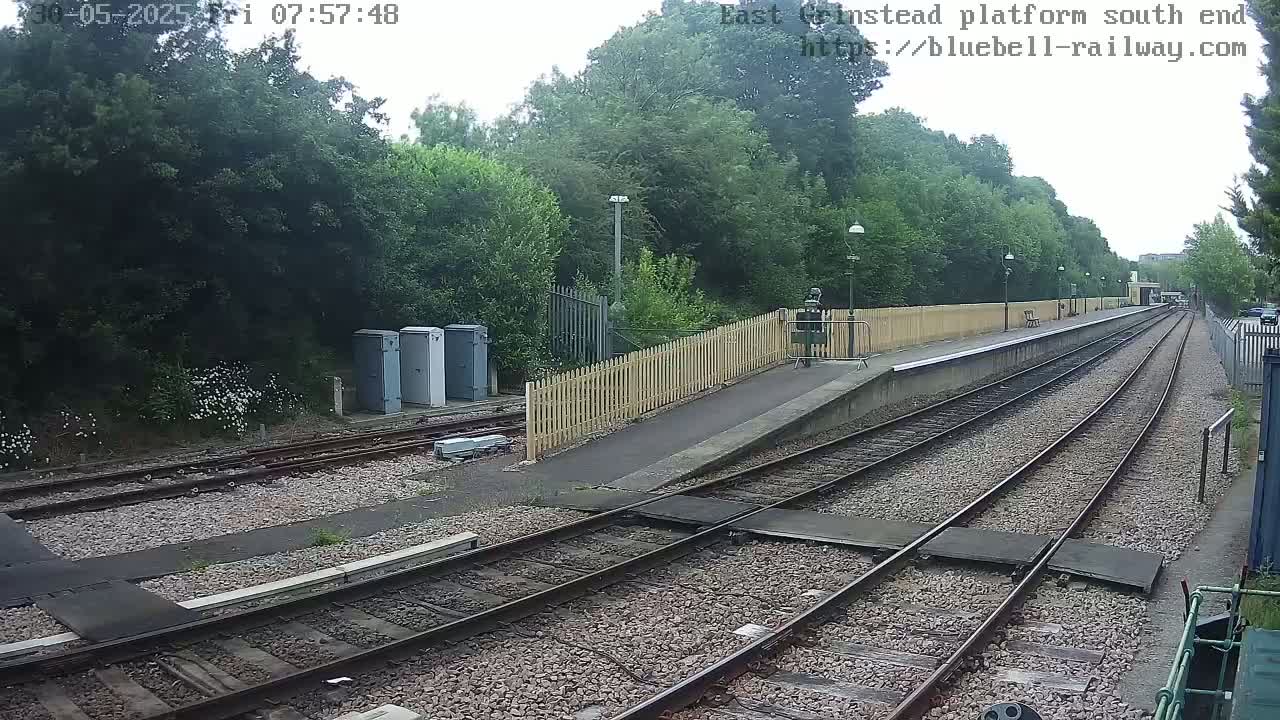 A view of a train platform on an overcast day, showing railway tracks extending into the distance, bordered by a wooden fence and lush green foliage.