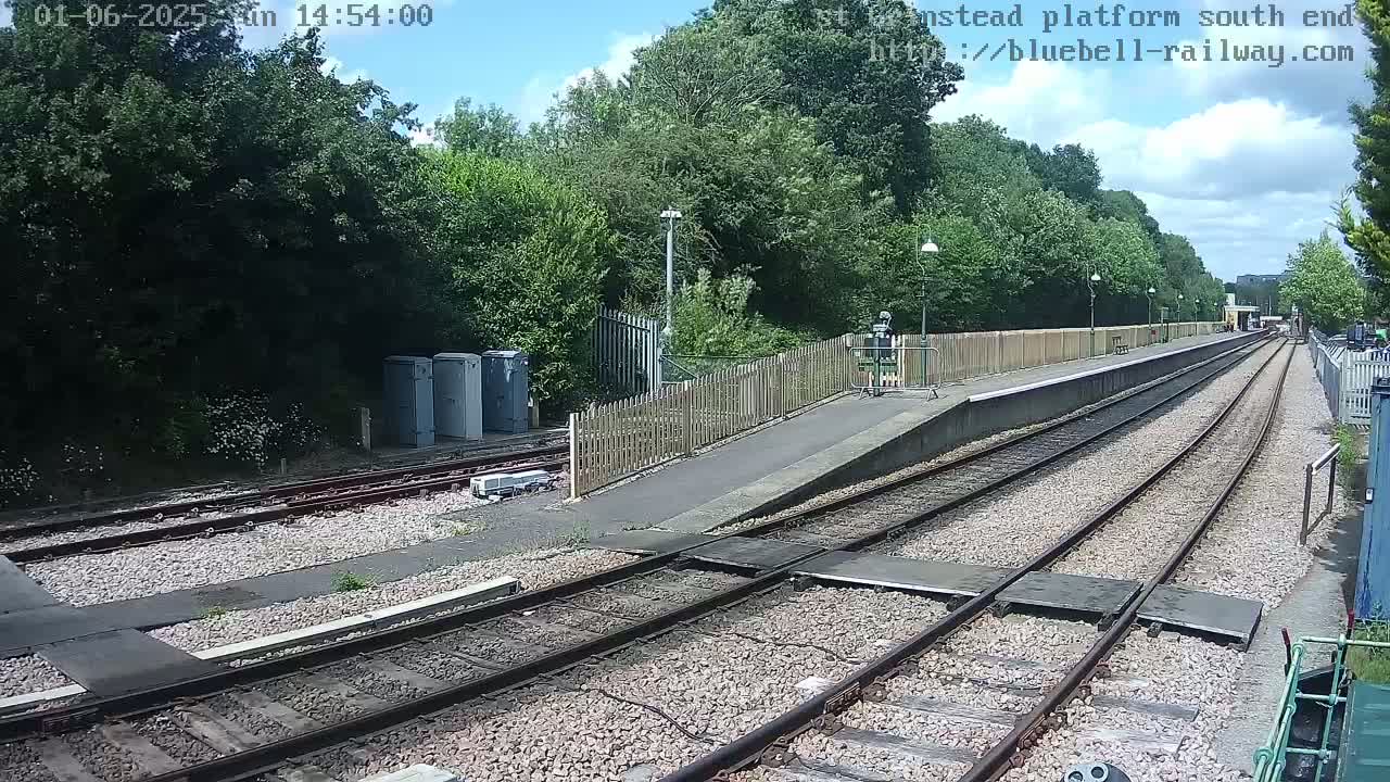 A train platform with two sets of tracks extends into the distance under a partly sunny sky, surrounded by lush green trees and a wooden fence.