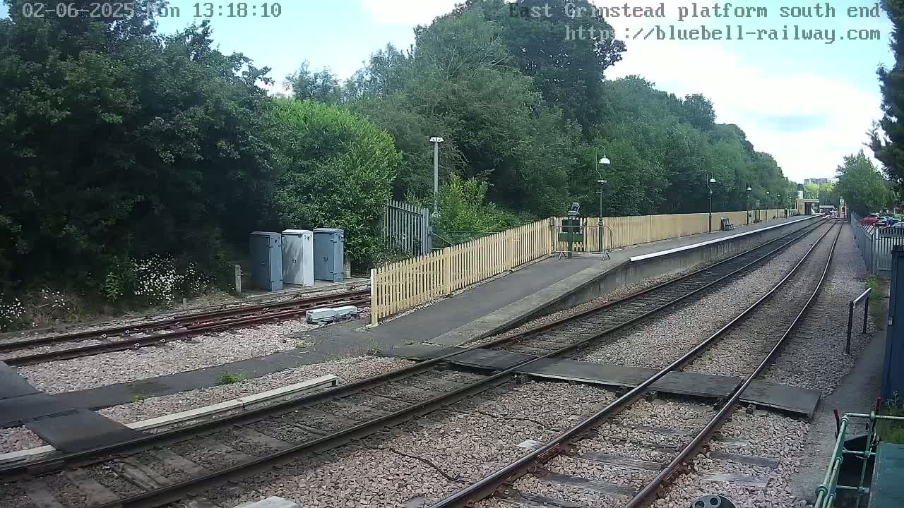 A train platform with two sets of tracks extends into a wooded area under a partly sunny sky.