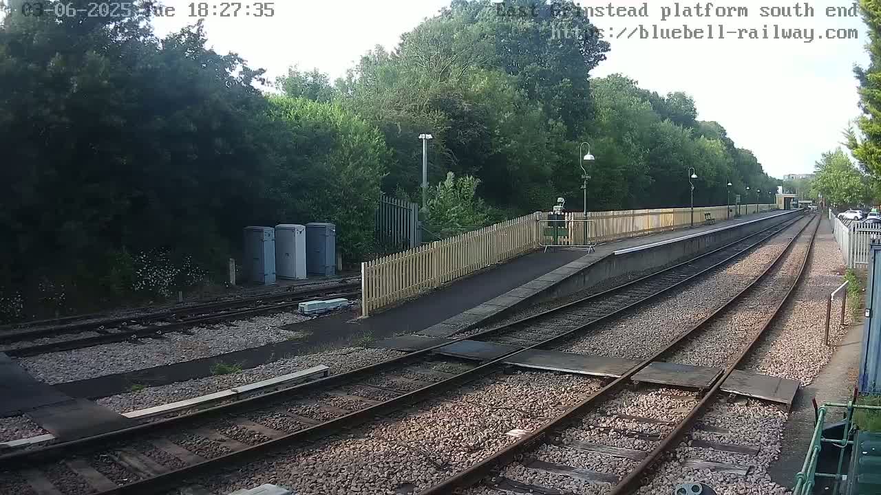 A train platform with multiple tracks extends into the distance, bordered by a wooden fence and lush greenery under a partly cloudy sky.
