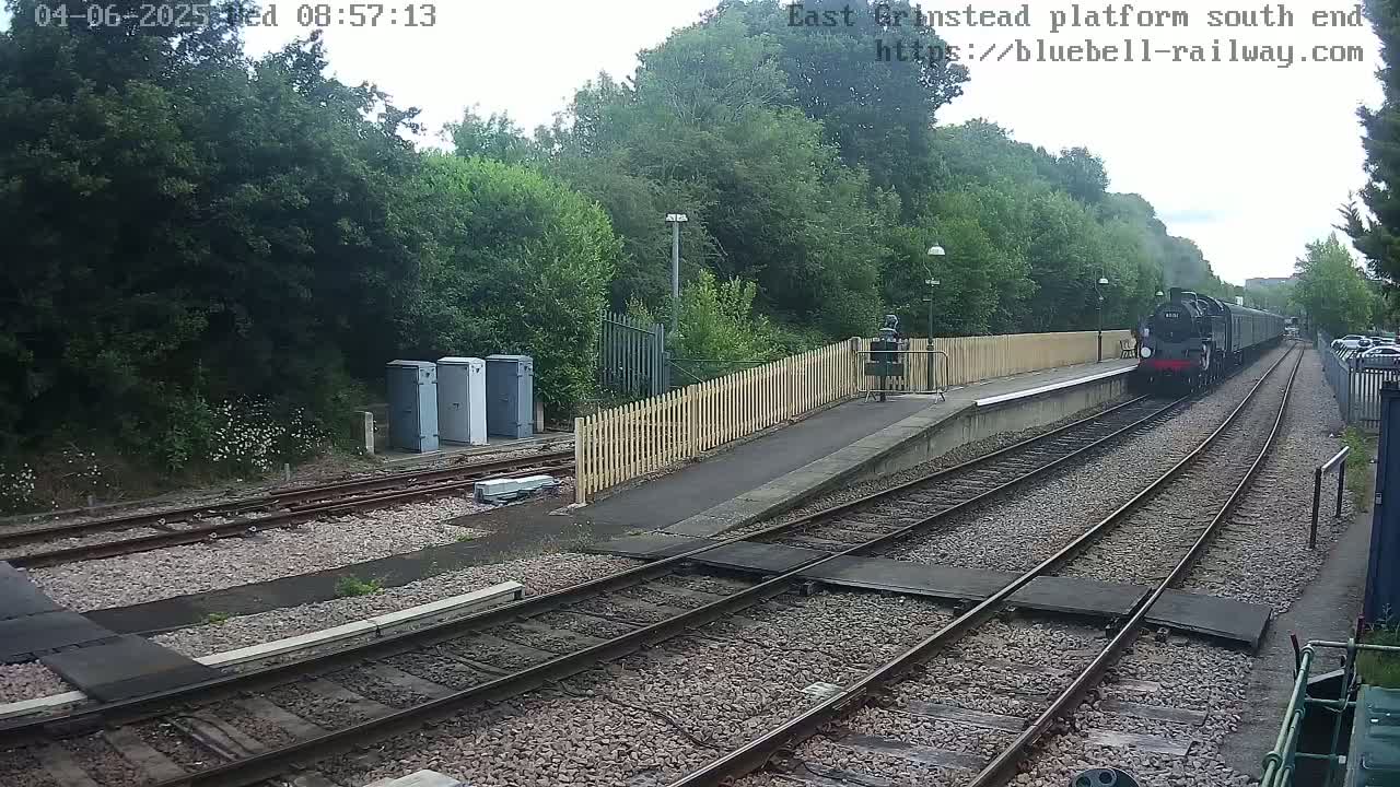 A steam train departs a railway station on an overcast day, passing alongside a wooden fence and a small platform.