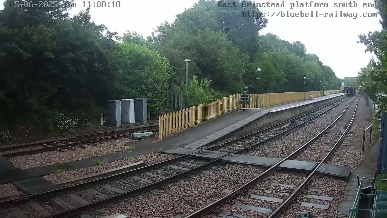 A train departs from a railway platform bordered by a wooden fence and lush green trees under an overcast sky.