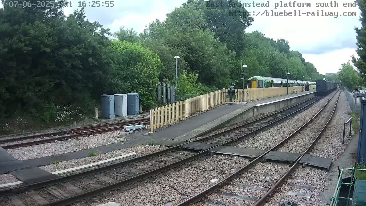 A train sits at a station platform next to a wooded area on a partly cloudy day.