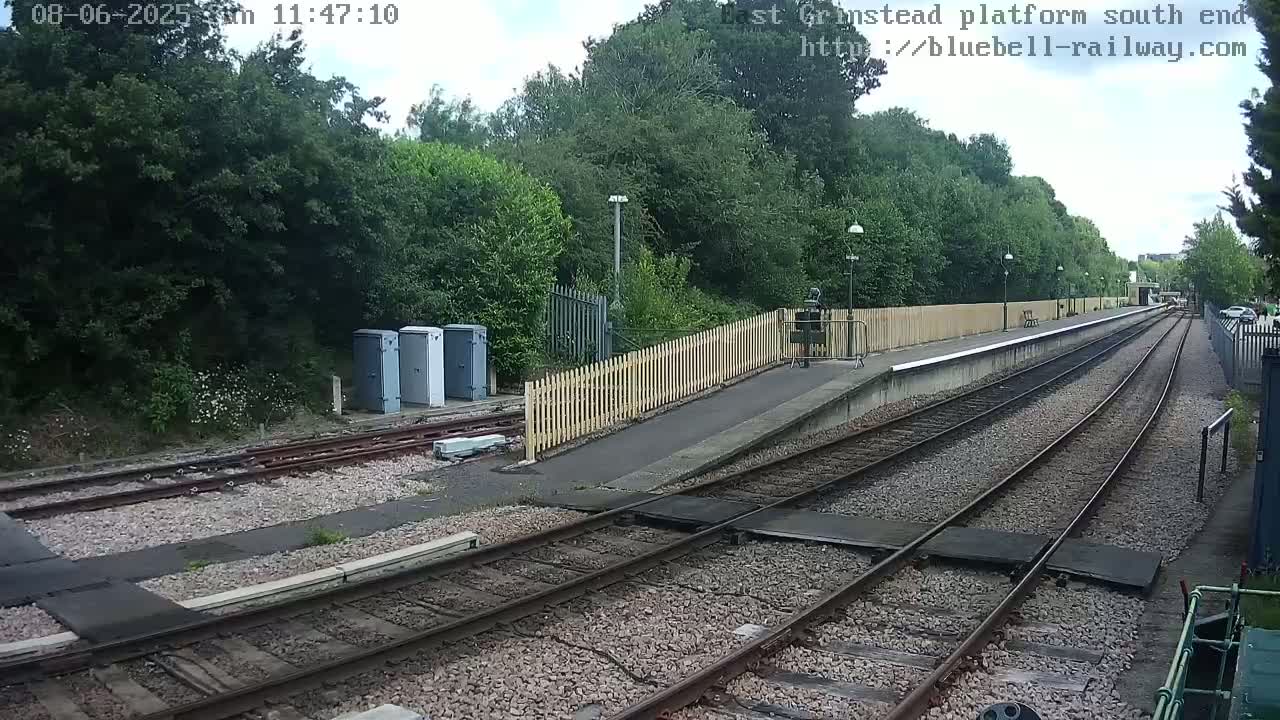 A train platform with two sets of tracks extends into the distance, bordered by a wooden fence and lush green trees under a partly cloudy sky.